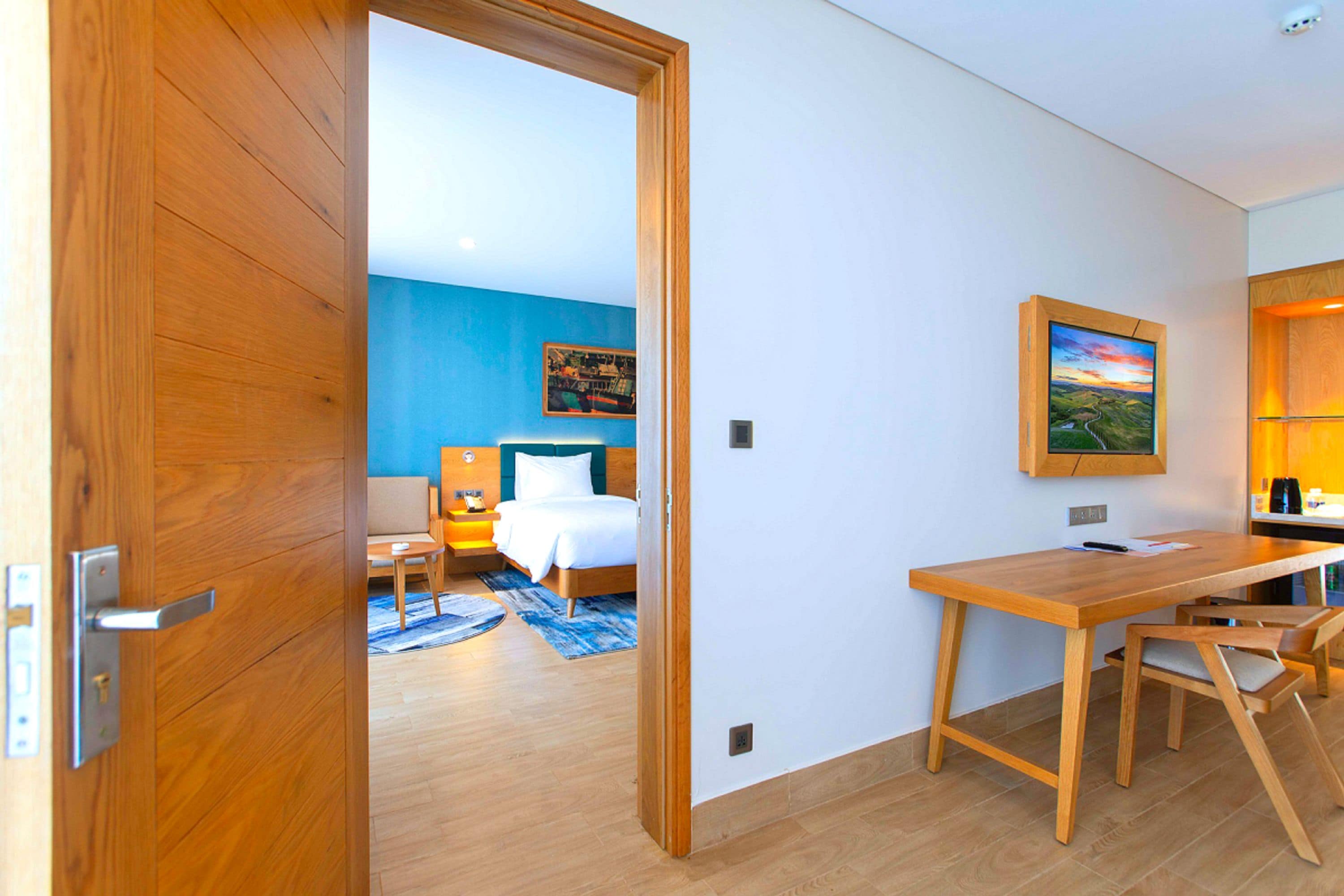A hotel room with a wooden door, a bed with white bedding, a desk with a chair, and a painting on the wall.