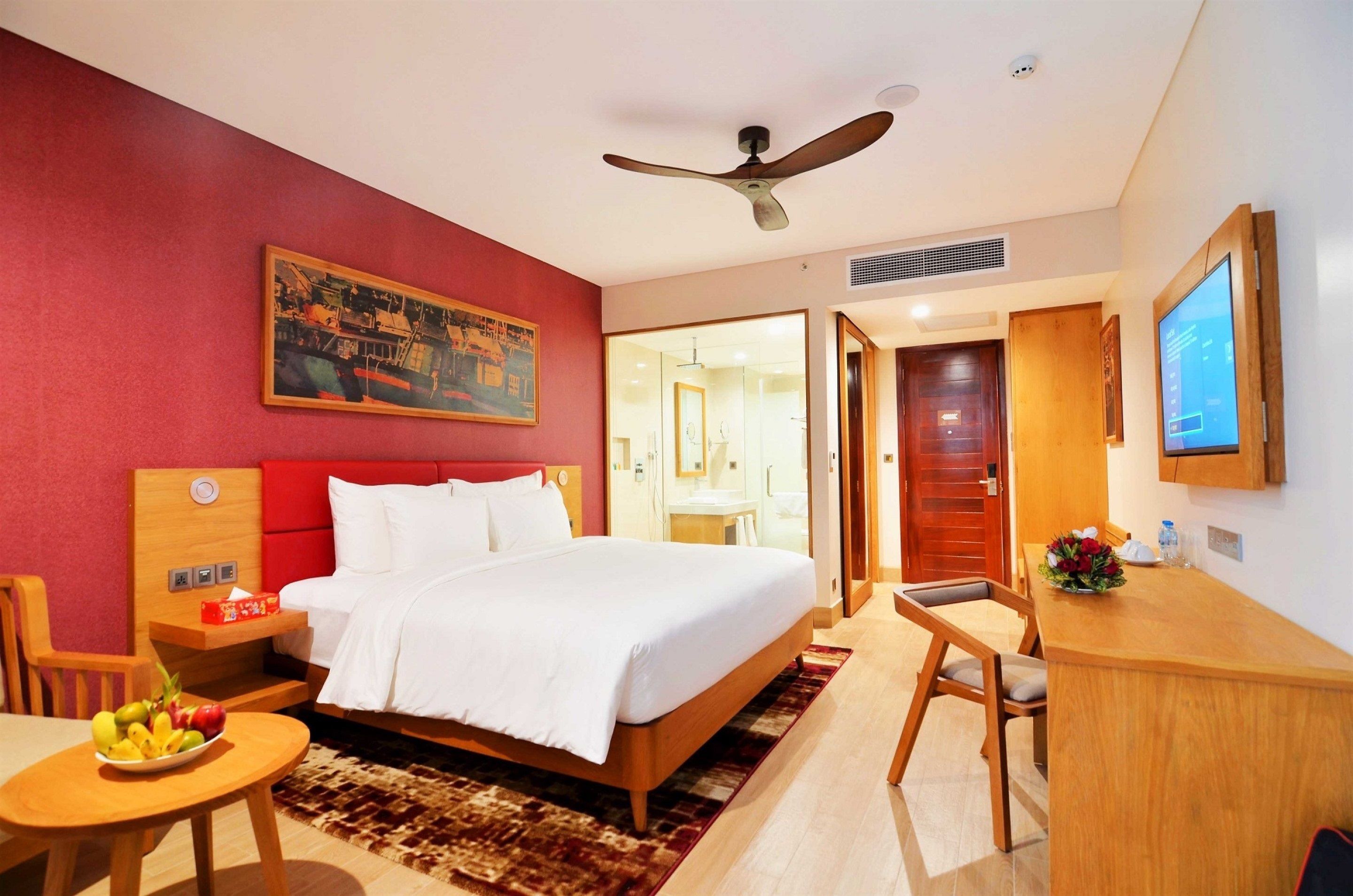 Habitación de hotel con cama, escritorio, silla, una mesita con fruta, un cuadro y televisor.