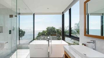 A modern bathroom with a large bathtub, a glass shower enclosure, and a view of the sea.