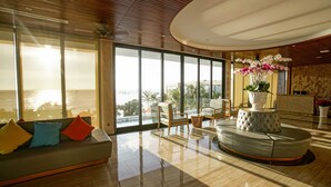 Een ruime woonkamer met een groot raam met uitzicht op zee, een moderne bank met kleurrijke kussens en een rond zitgedeelte met een decoratief middelpunt.