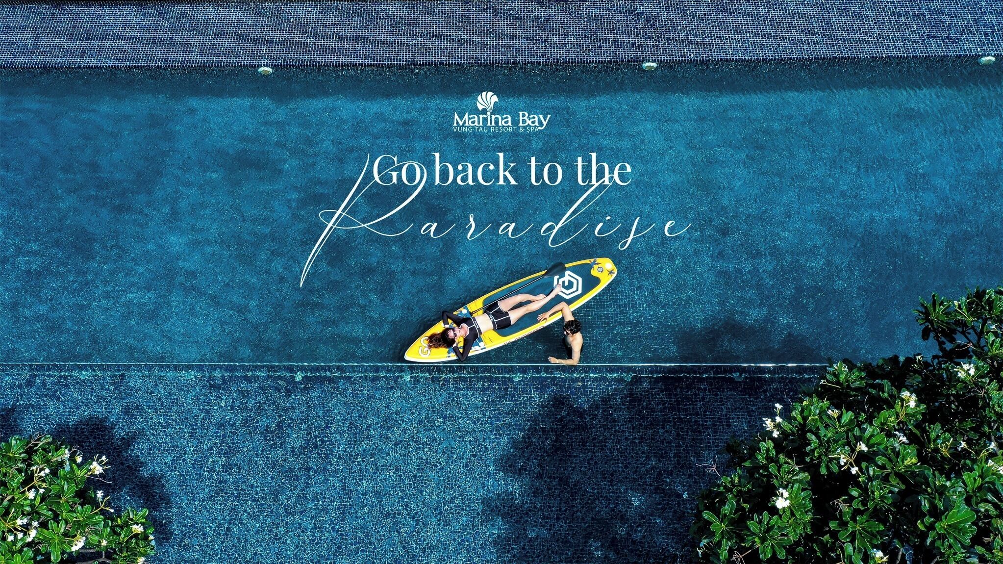 A swimming pool with a person lying on a yellow surfboard, surrounded by greenery.