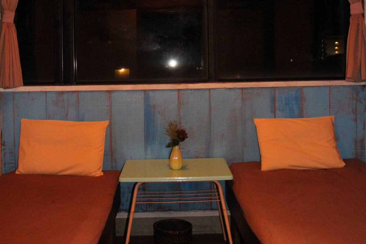 Ein Zimmer mit zwei Betten, einem kleinen Tisch mit einer Vase und einem Fenster mit Vorhängen.