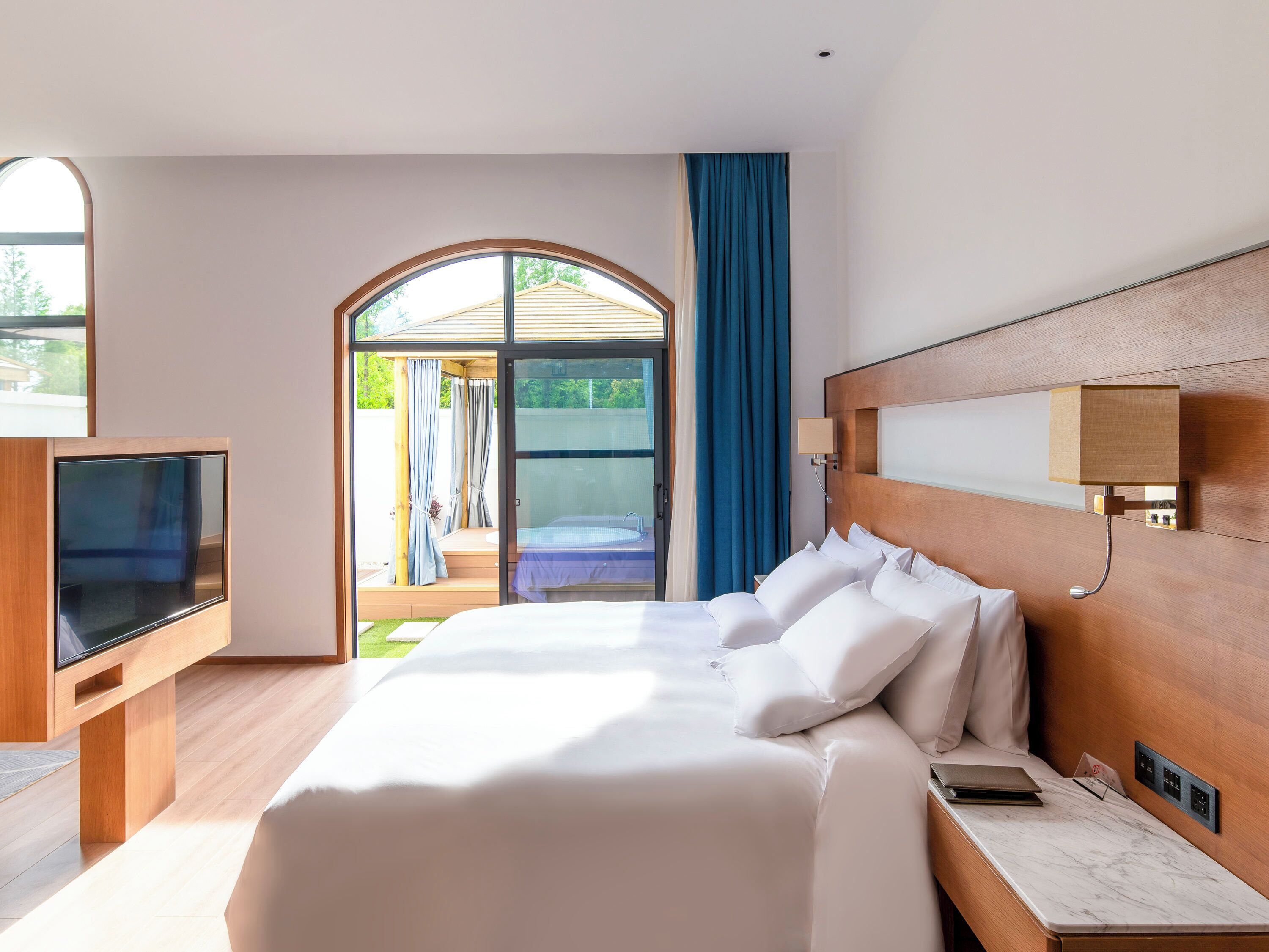 A hotel room with a large bed, a TV, a marble bedside table, and a view of the outdoors.