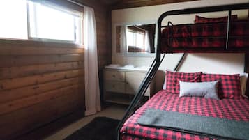 Un lit superposé avec une couverture à carreaux, une table de chevet avec un miroir et une commode en bois.