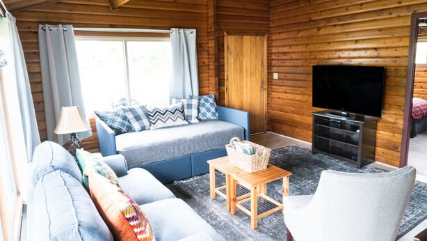A cozy cabin interior with a blue sofa, a wooden table, a television, and a window with curtains.