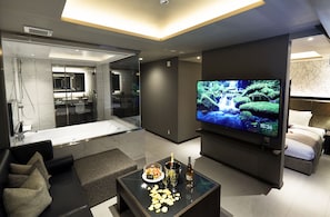 A modern living room with a flat-screen TV, a sofa, a coffee table with snacks and drinks, and a glass-enclosed bathroom. - Bamboo Garden Tokyo - Adults Only (Tokyo)