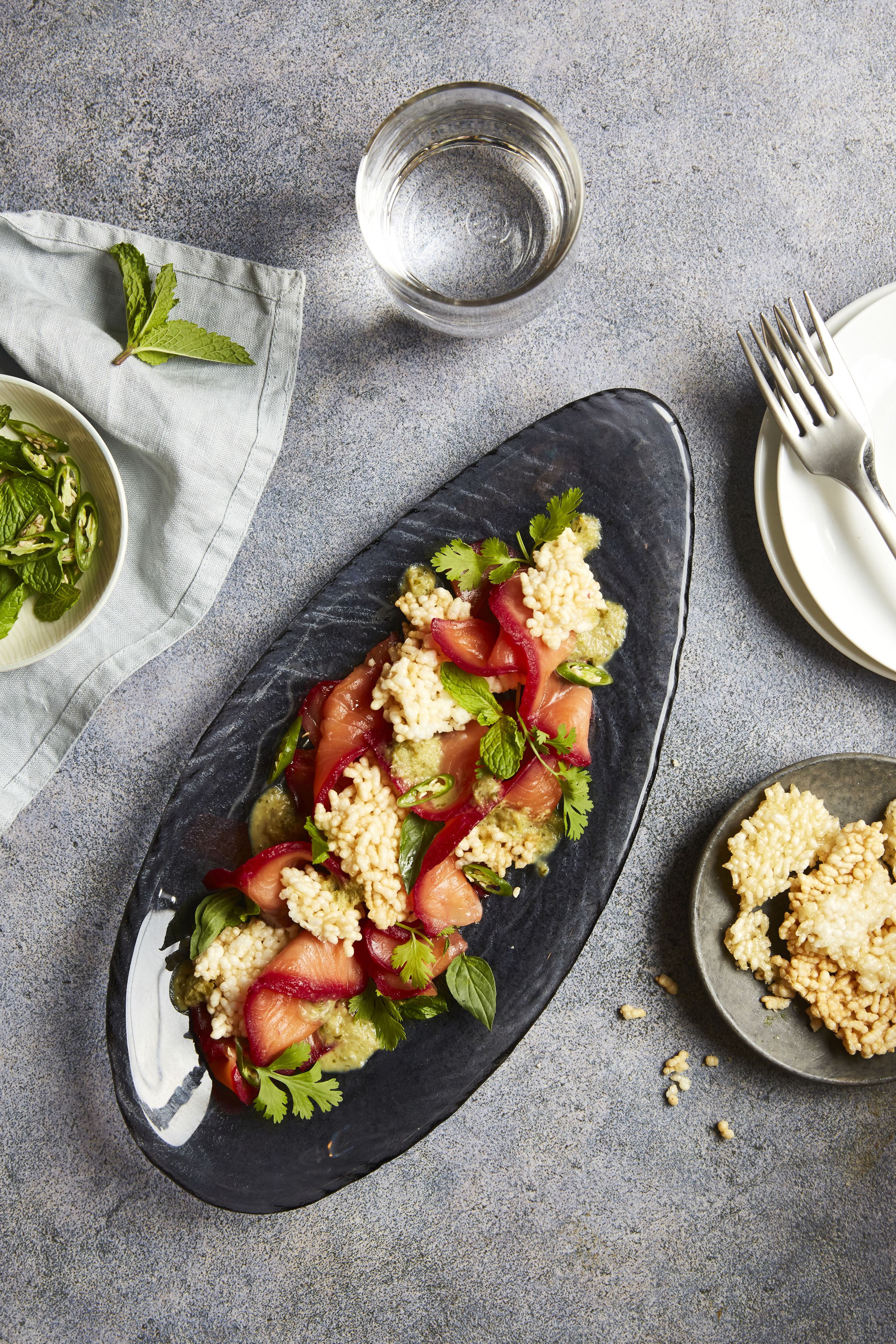 Ein dunkler Teller mit einem Salat aus Tomaten, Gurken und Kräutern auf einer strukturierten, grauen Oberfläche.