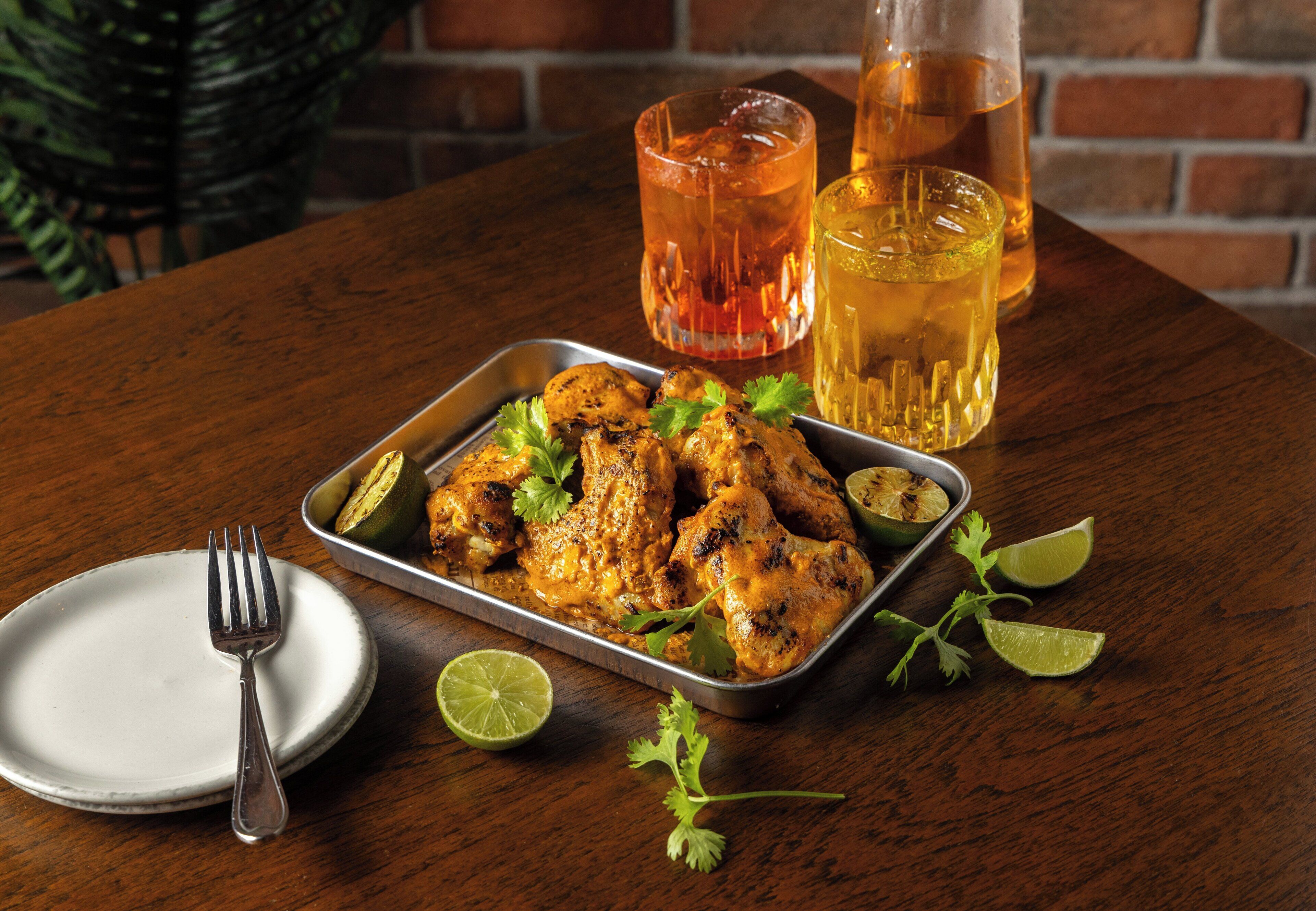 A tray of grilled chicken with lime wedges and cilantro, a glass of iced tea, and a bottle of water on a wooden table.