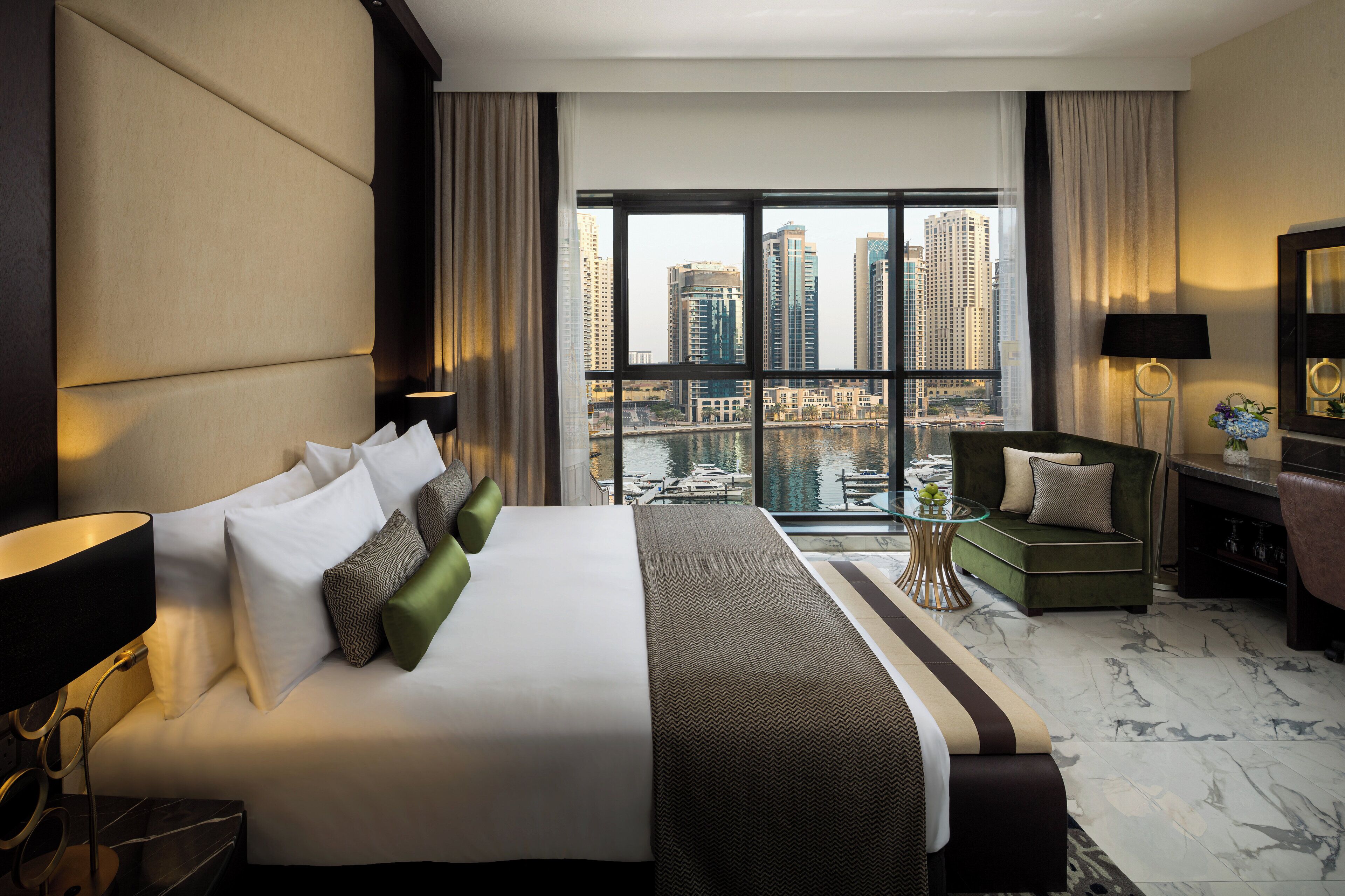 A hotel room with a large bed, a sofa, a glass table, and a view of a cityscape through large windows.