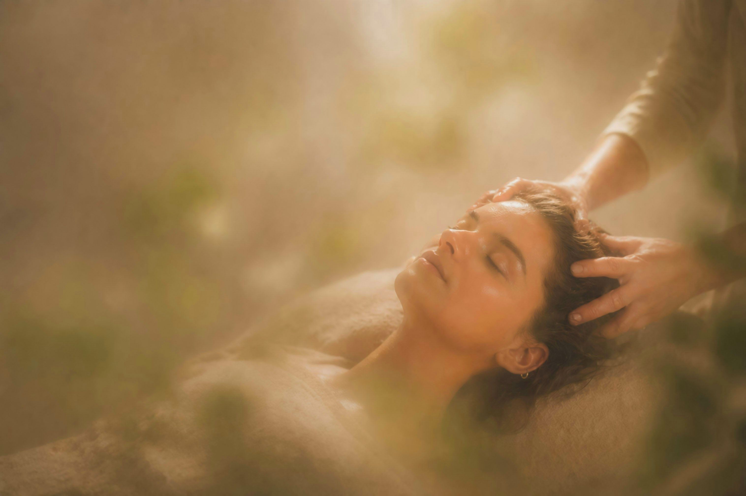 A person lying down with a hand gently touching their head, in a room with soft lighting and a blurred background.