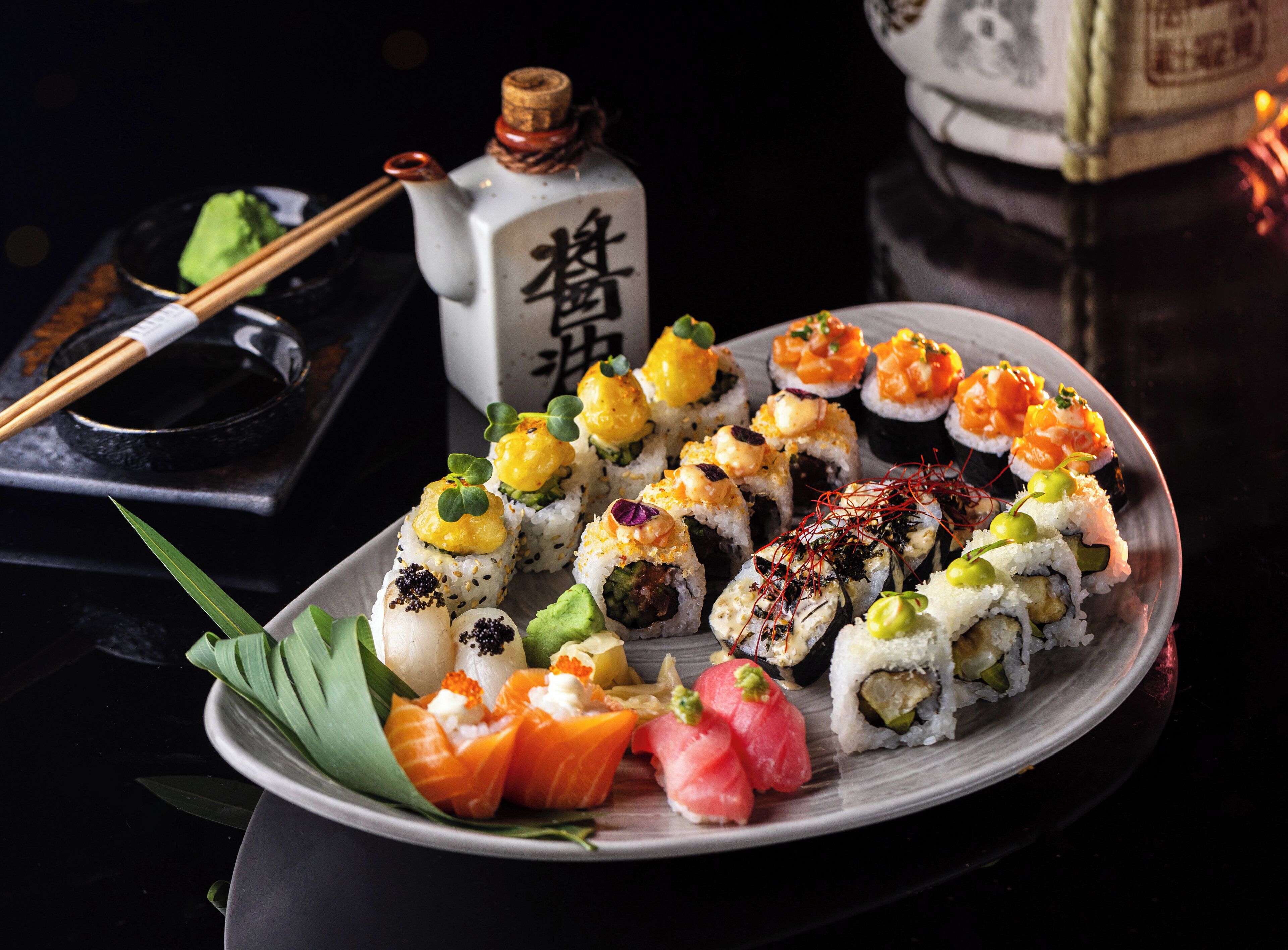 A variety of sushi on a plate, a bottle with Japanese characters, chopsticks, and a small bowl.