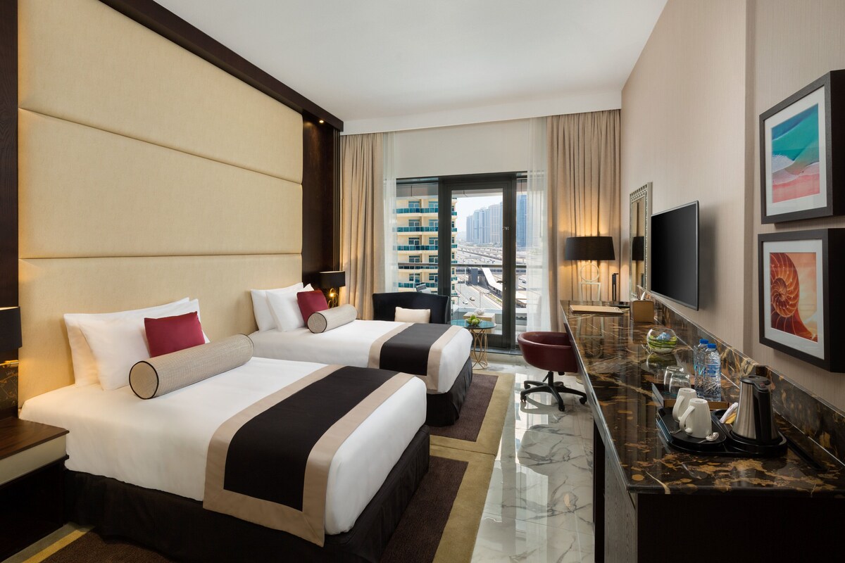 A hotel room with two beds, a desk with a coffee maker, a TV, and a view of the city.