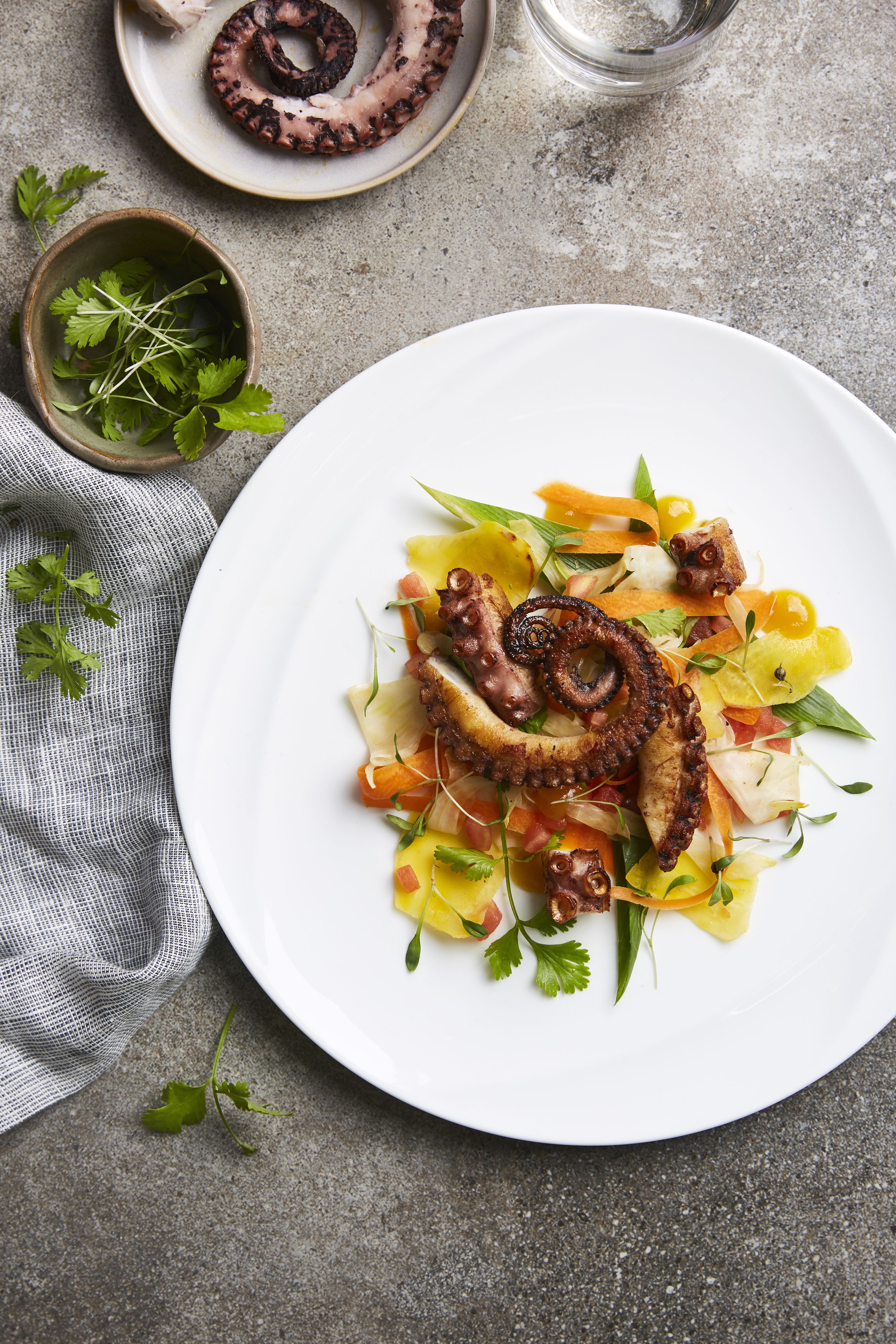 A plate of octopus salad with a bowl of greens and a glass of water on a textured surface.