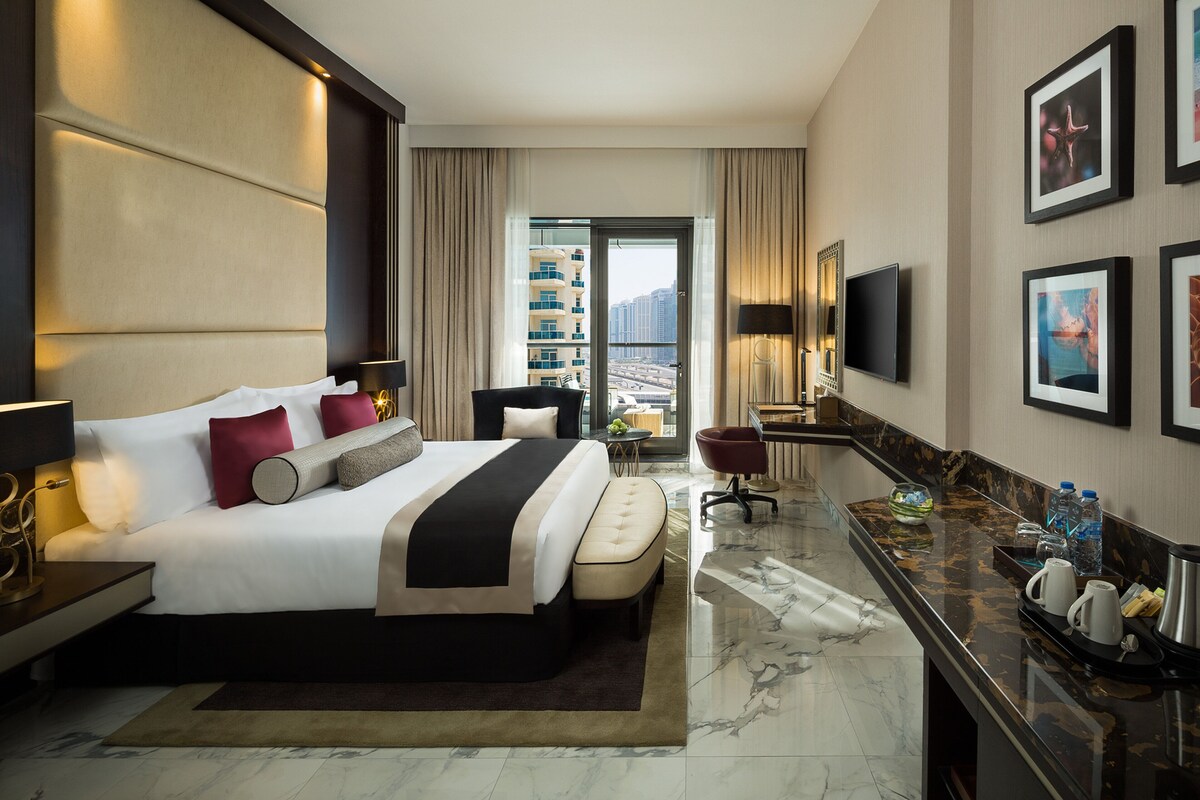 A modern hotel room with a large bed, a desk, a chair, a TV, and a view of the city.