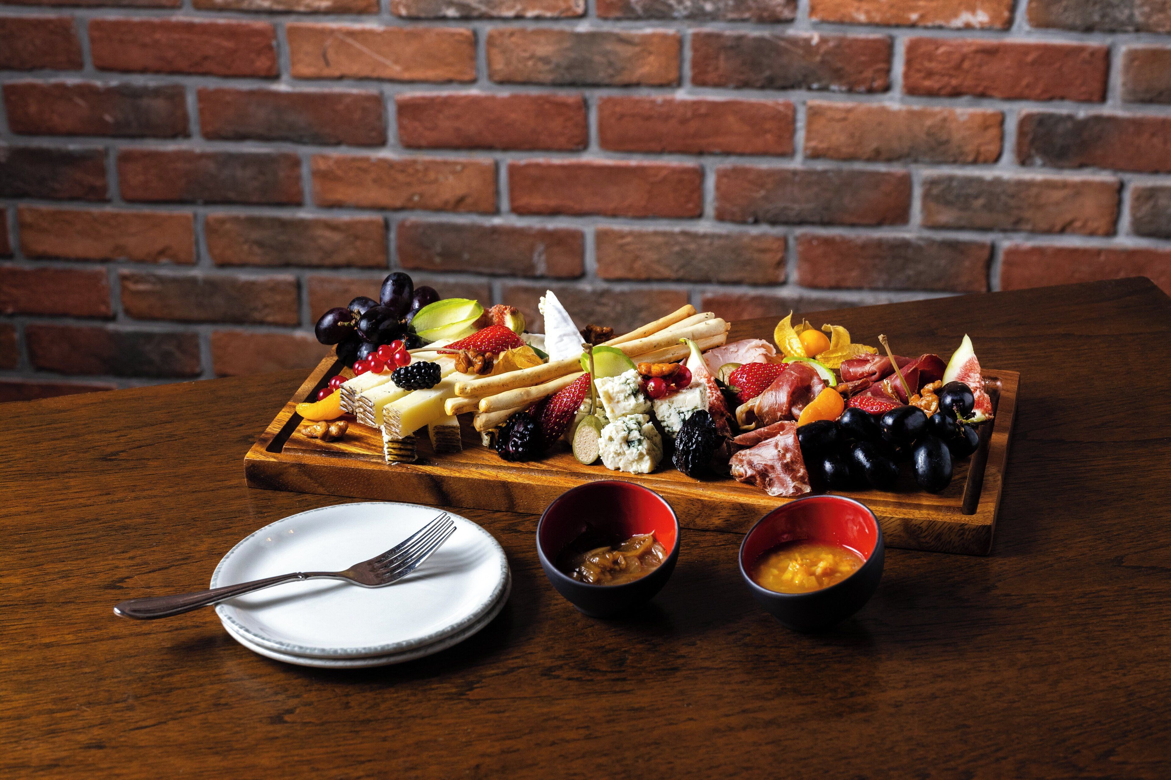 Une planche en bois sur laquelle sont disposés un assortiment de fromages, de fruits et de charcuteries, le tout posé sur une table devant un mur en briques.
