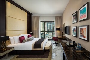 A modern hotel room with a large bed, a desk, a chair, a TV, and a view of the city.