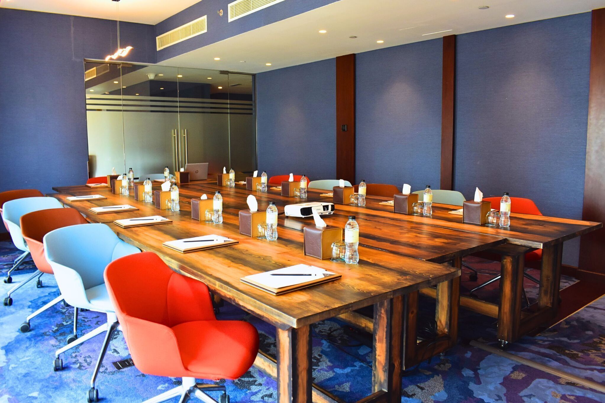 Un salón de conferencias con una larga mesa de madera, sillas coloridas y botellas de agua sobre la mesa.