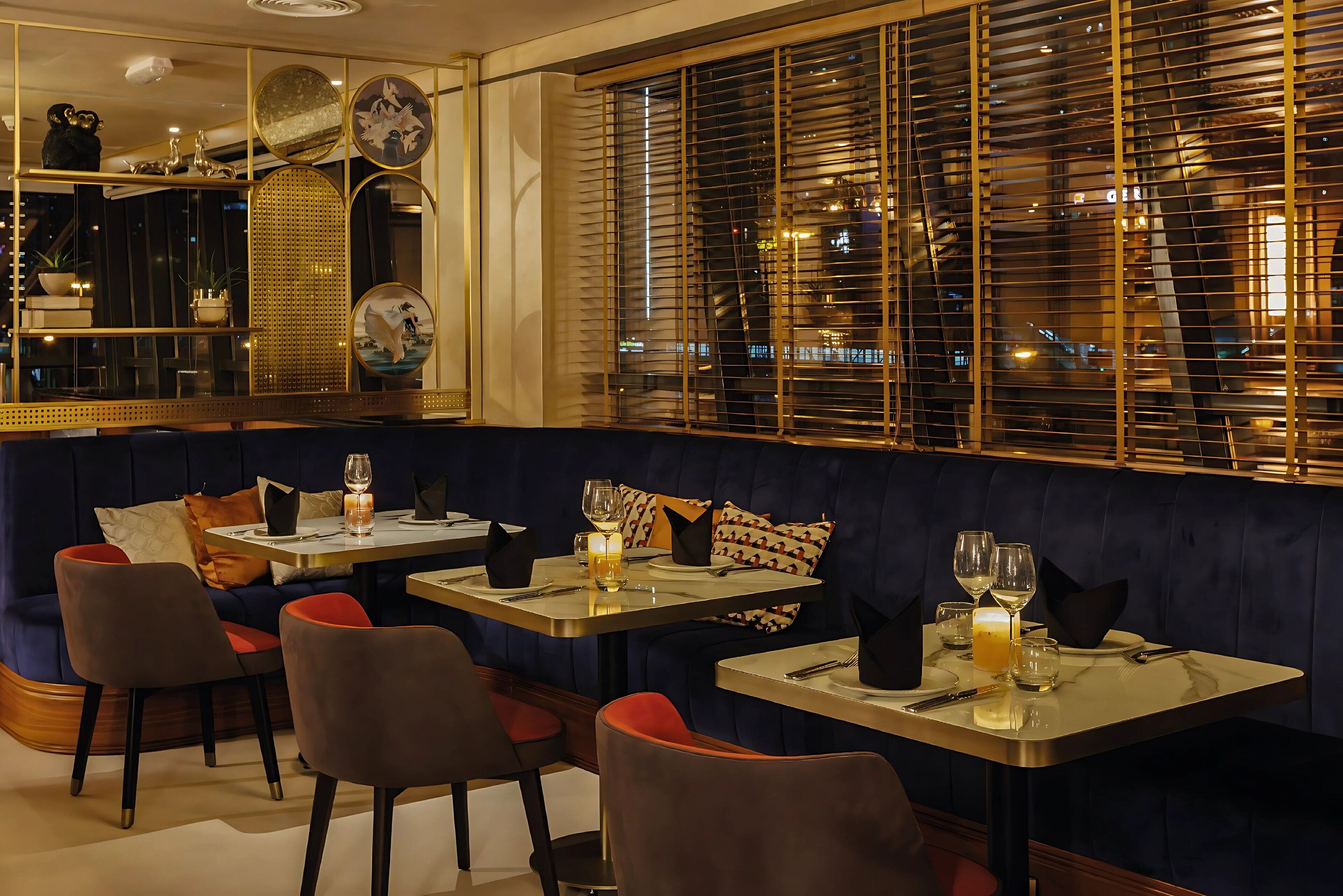 Ein Restaurant in gedämpfter Beleuchtung mit Sitzecken und Tischen, das einen Blick auf die Stadt durch große Fenster mit Jalousien bietet.