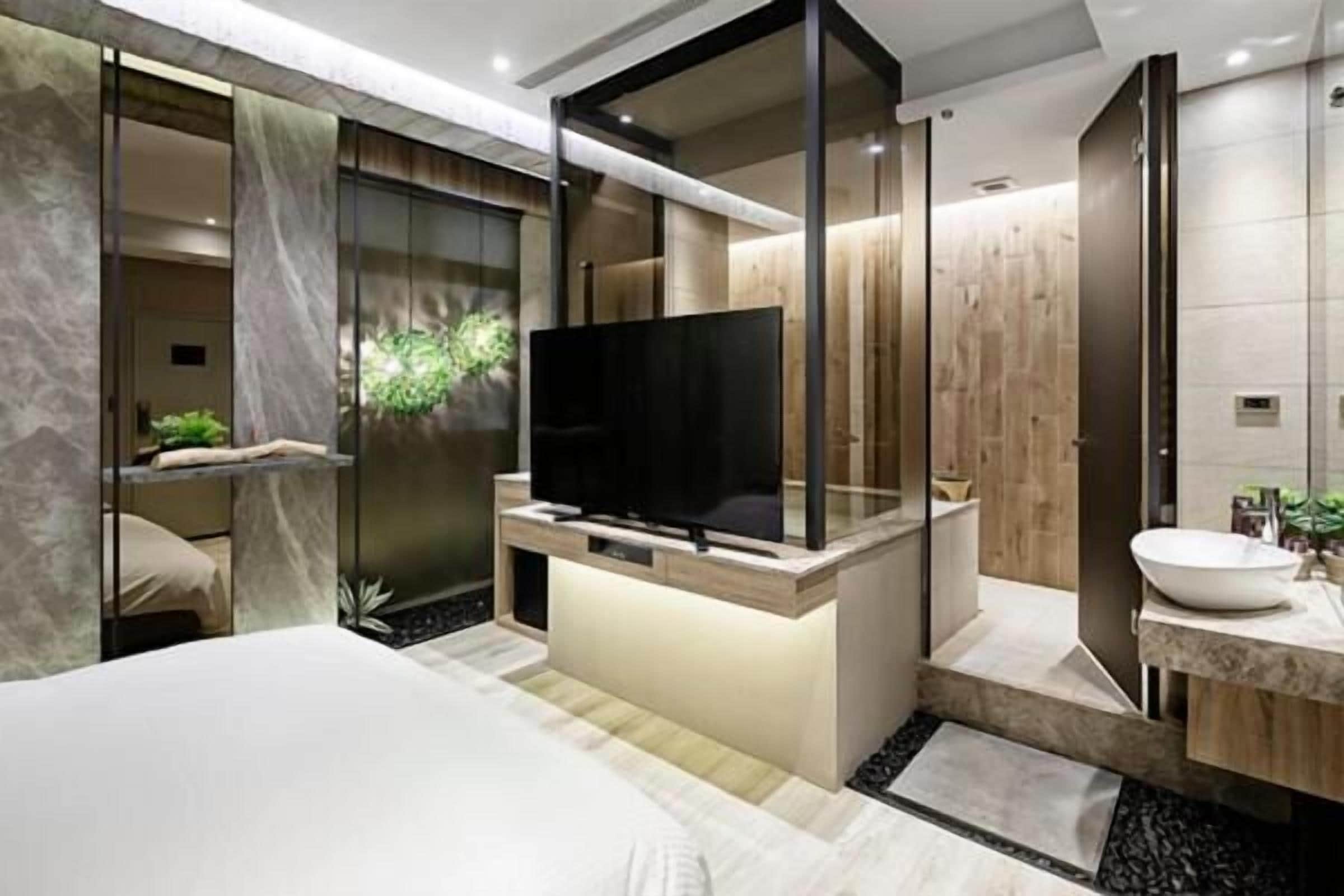 A modern hotel room with a large bed, a television, a bathroom with a sink and mirror, and a small plant.