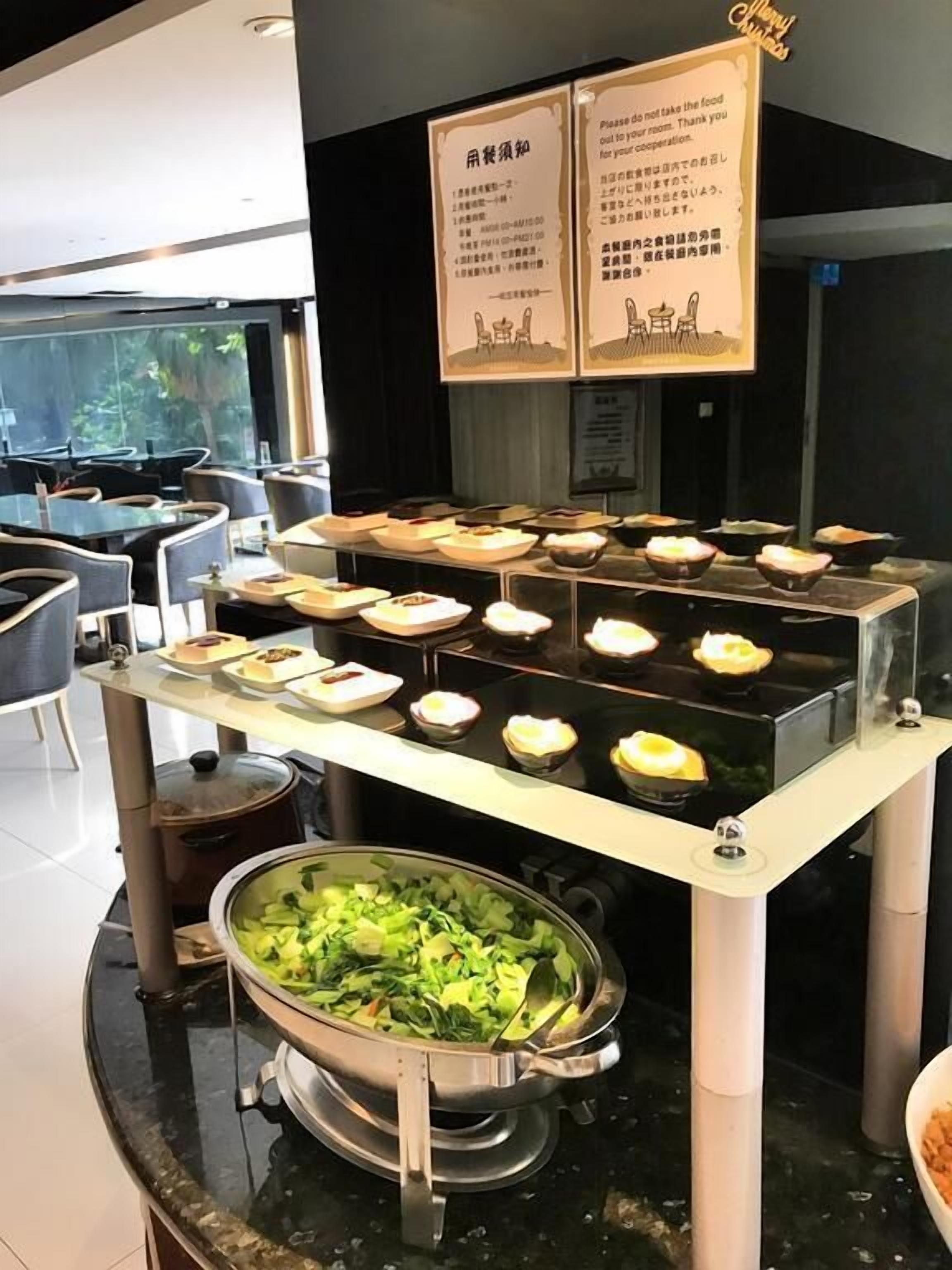 Uma área de buffet com diversos pratos expostos em um balcão, um caldeirão de legumes e duas placas informativas.