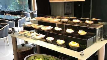 Uma área de buffet com diversos pratos expostos em um balcão, um caldeirão de legumes e duas placas informativas.