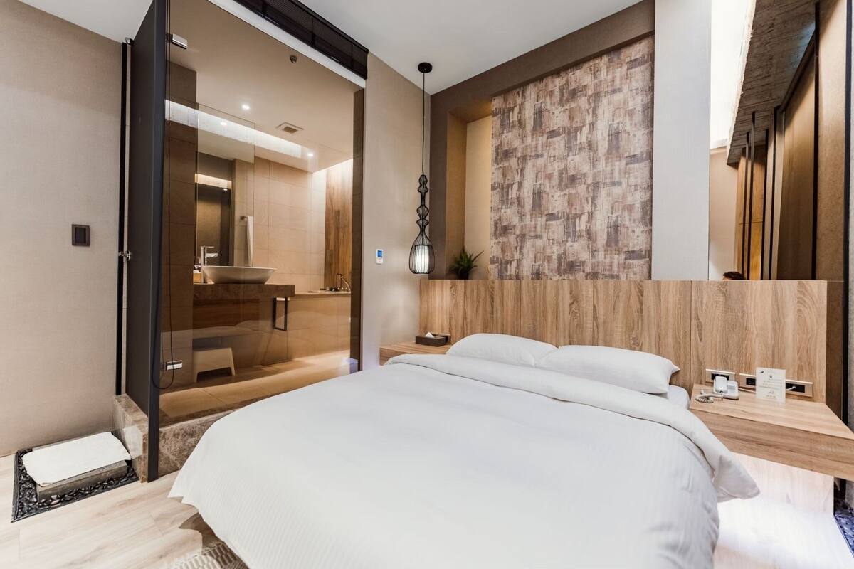 A modern hotel room with a large bed, a wooden headboard, a bedside table with a lamp, and a view of the bathroom.