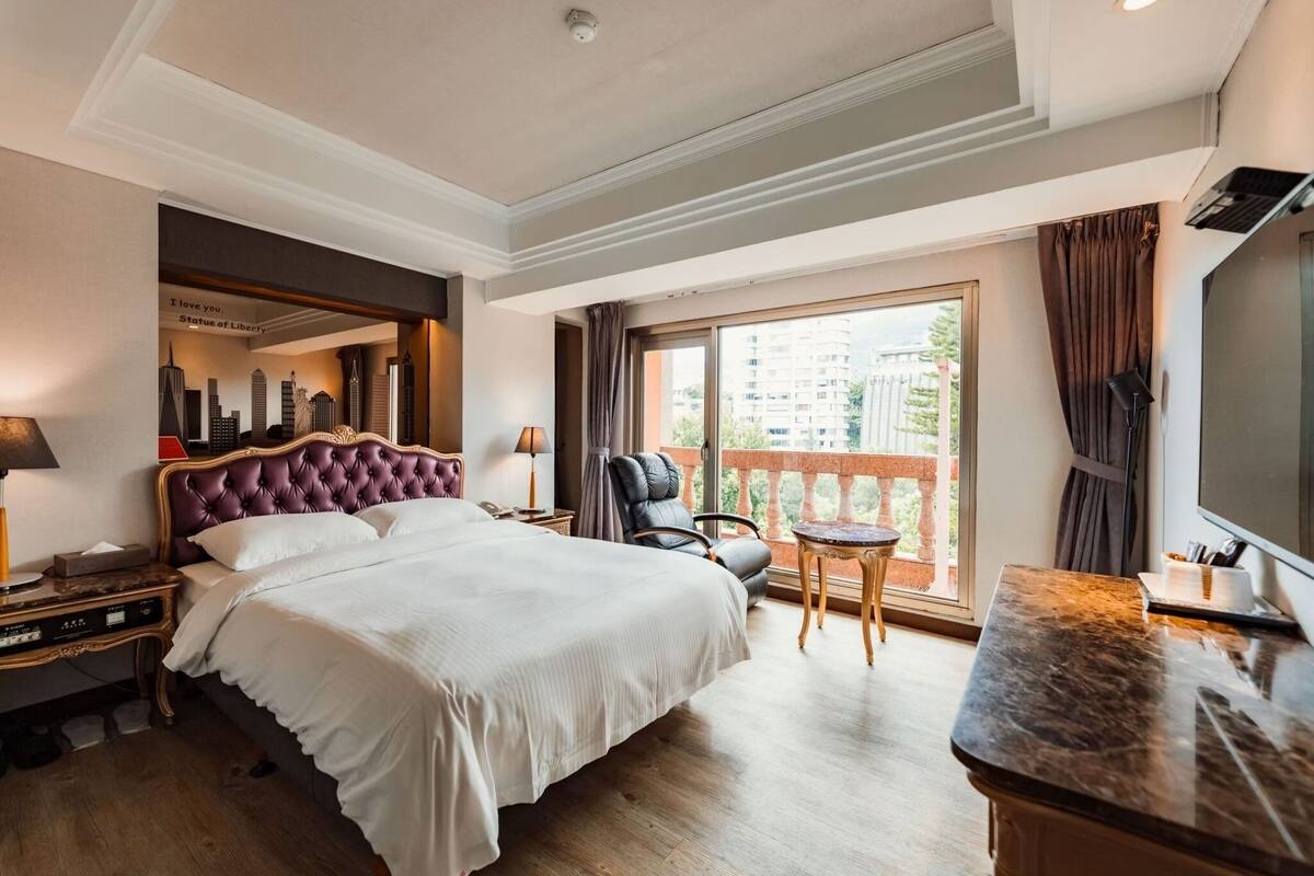 A hotel room with a large bed, a desk, a chair, a TV, and a balcony with a city view.