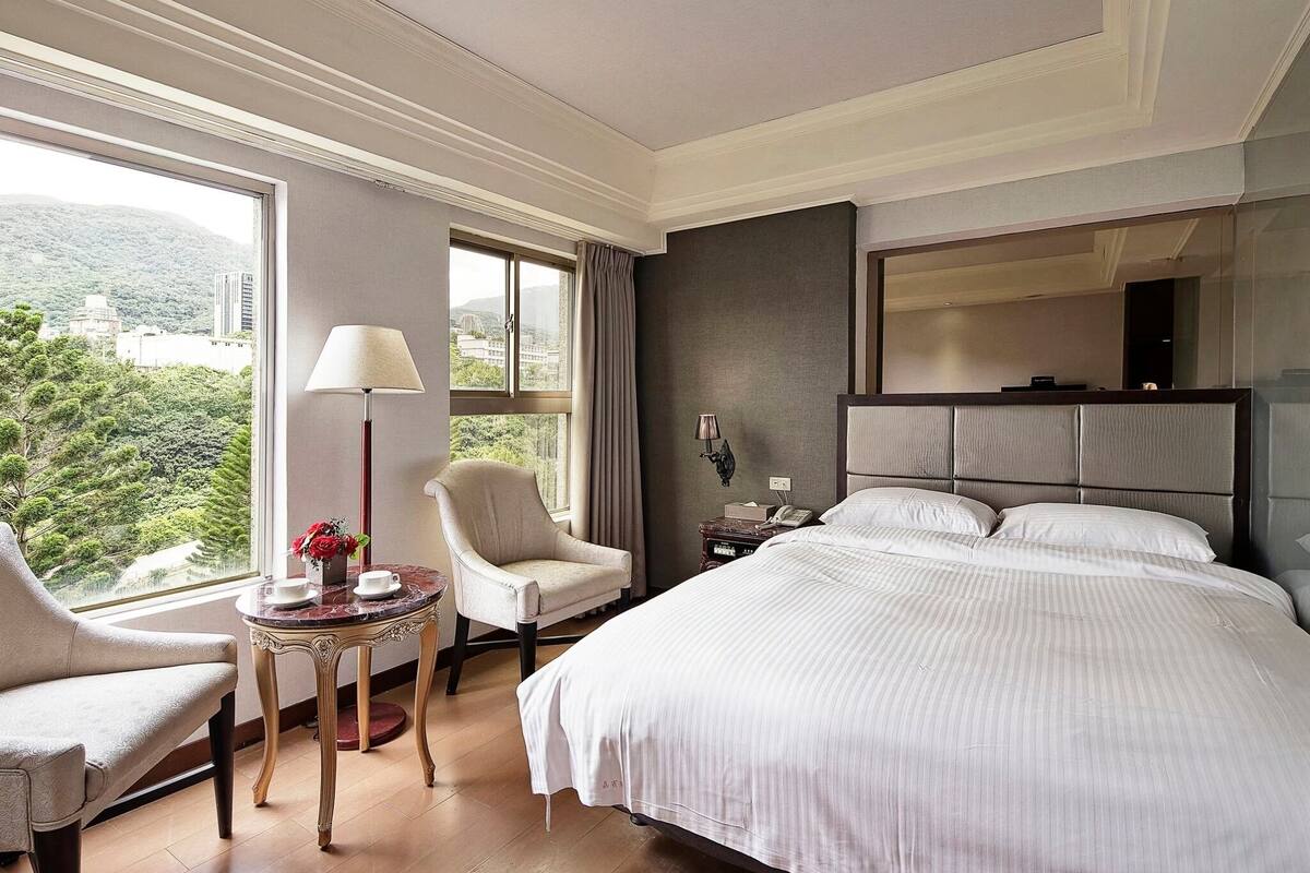 A hotel room with a large bed, a chair, a small table with a lamp, a nightstand, and a window with a view of greenery.