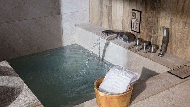 Une baignoire remplie d’eau, un seau en bois avec une serviette, et un mur recouvert de panneaux en bois.