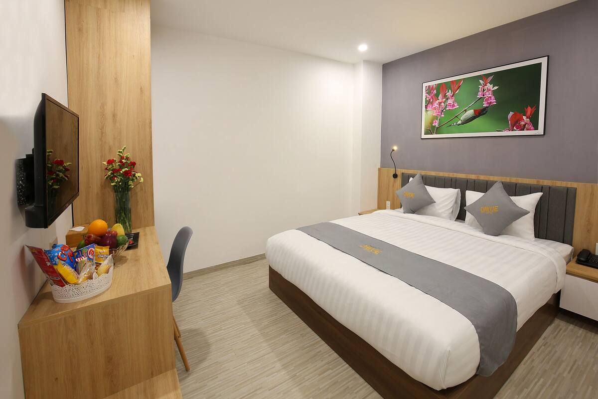 Quarto de hotel com cama, escrivaninha com cesta de frutas, cadeira, televisão e um quadro na parede.