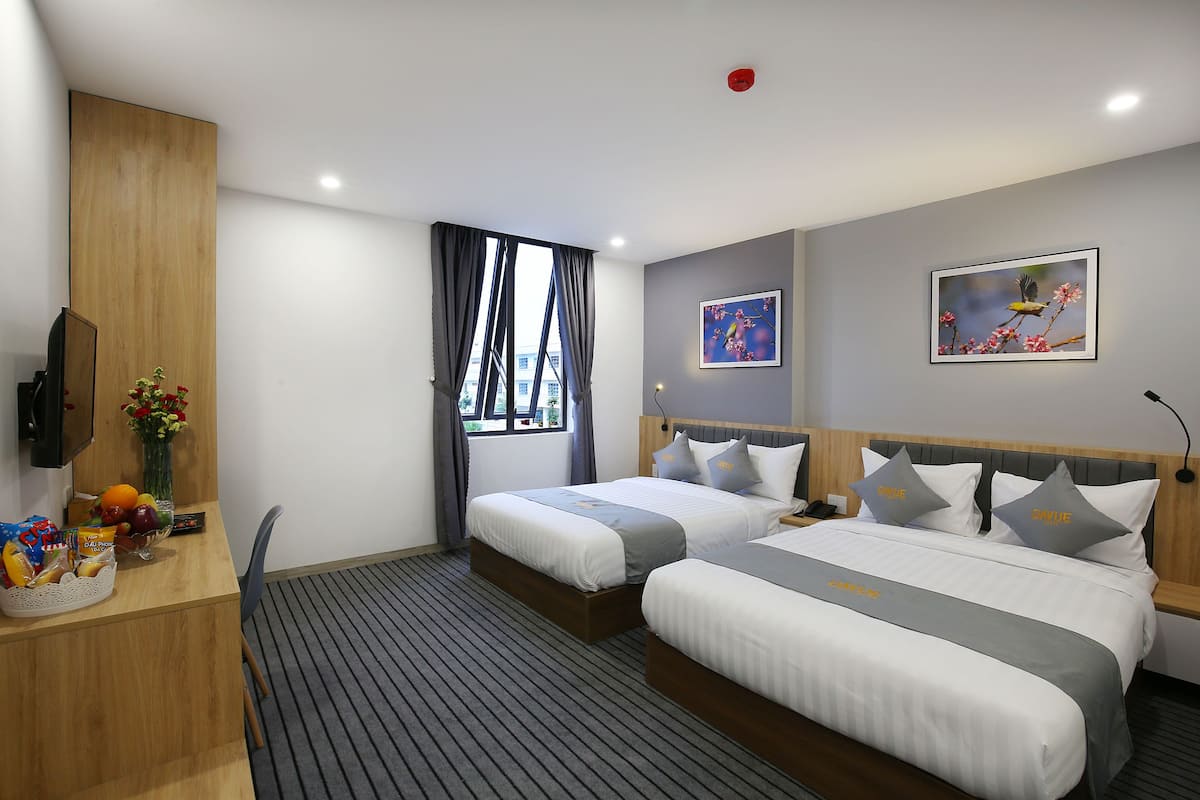 Quarto de hotel com duas camas, uma escrivaninha com um vaso de flores e uma janela com cortinas.