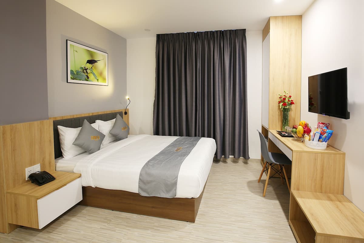 Quarto de hotel com cama, mesa de cabeceira com telefone, escrivaninha com cadeira, televisão e uma janela grande com cortinas.
