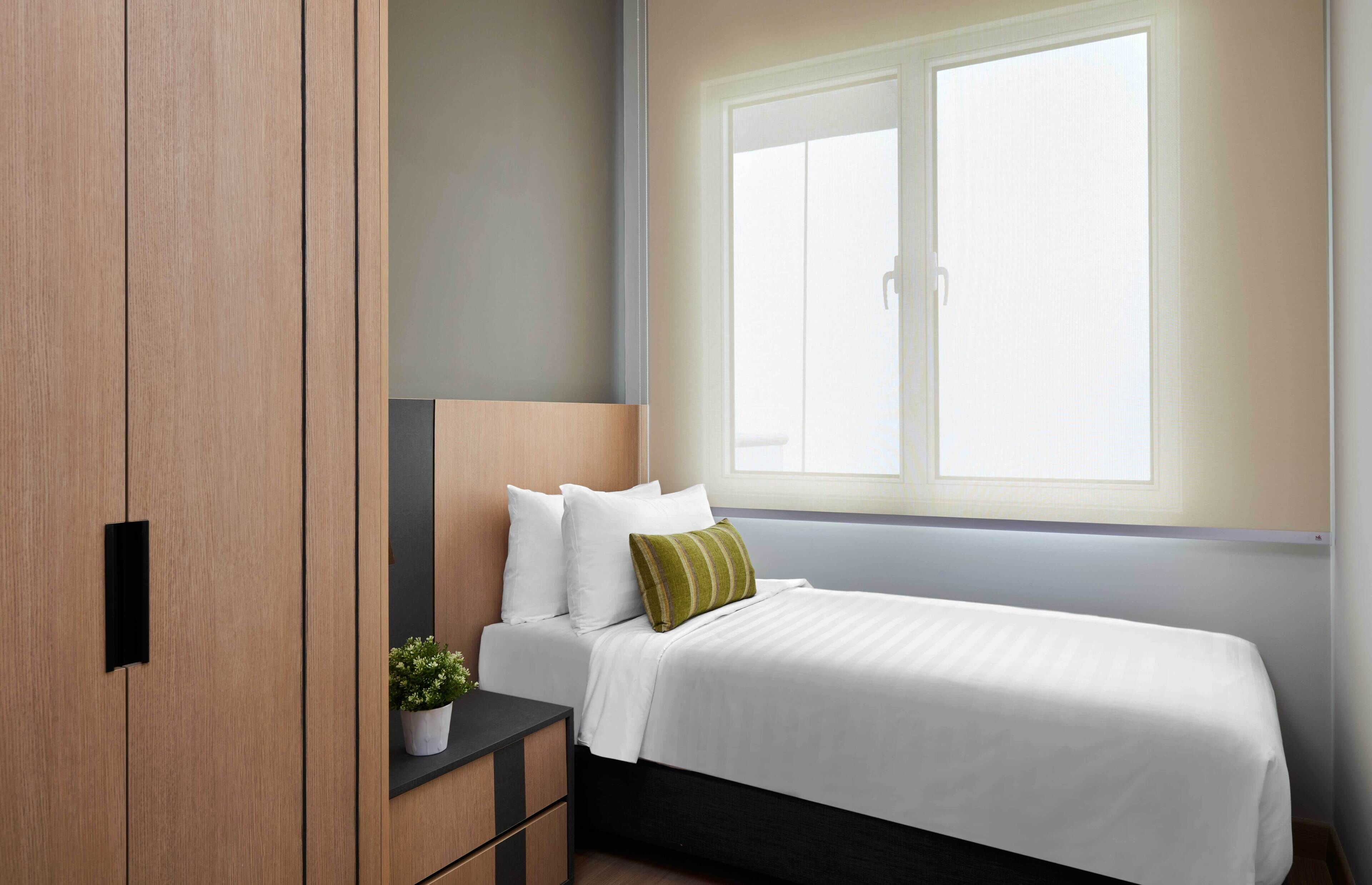 Un dormitorio con cama, mesita de noche, planta en maceta y ventana con persianas.