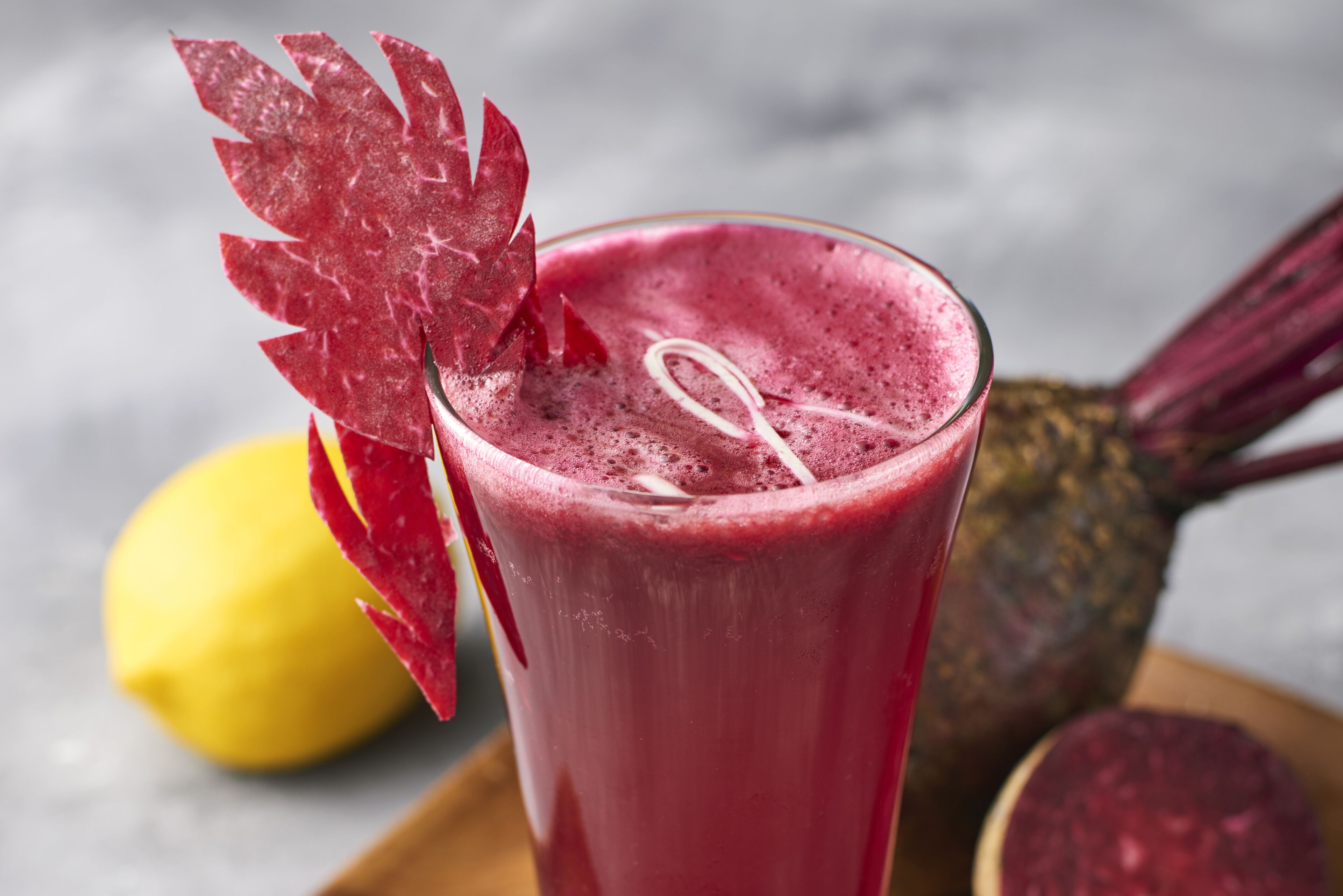 Un verre de smoothie rouge, décoré d’une feuille, d’un quartier de citron et d’un ananas en arrière-plan.