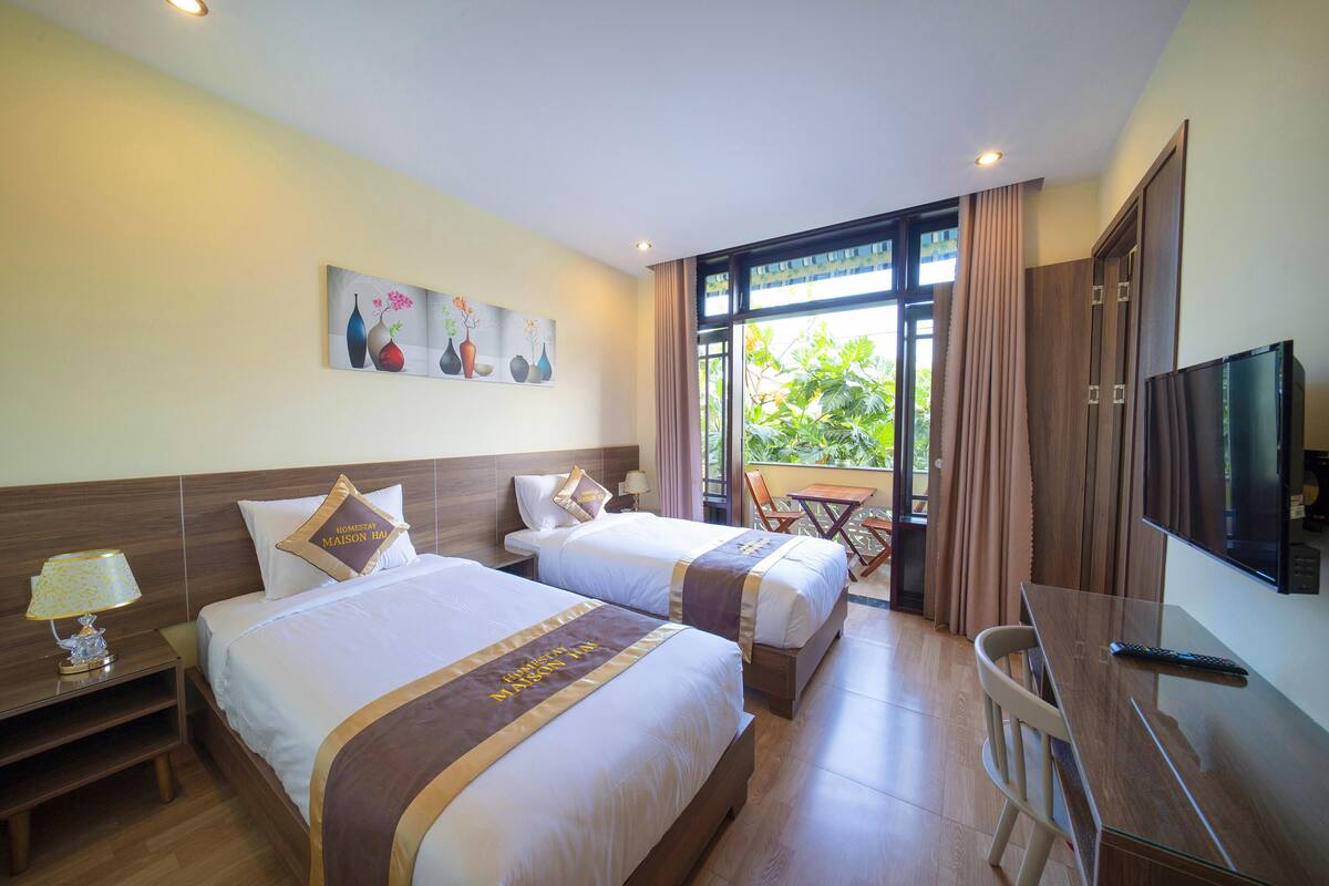 A hotel room with two beds, a TV, a desk, and a balcony with curtains.