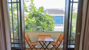 A balcony with a wooden table and two chairs, overlooking a garden with large leafy plants and a blue building in the background. - Maison Hai Homestay (Da Nang)