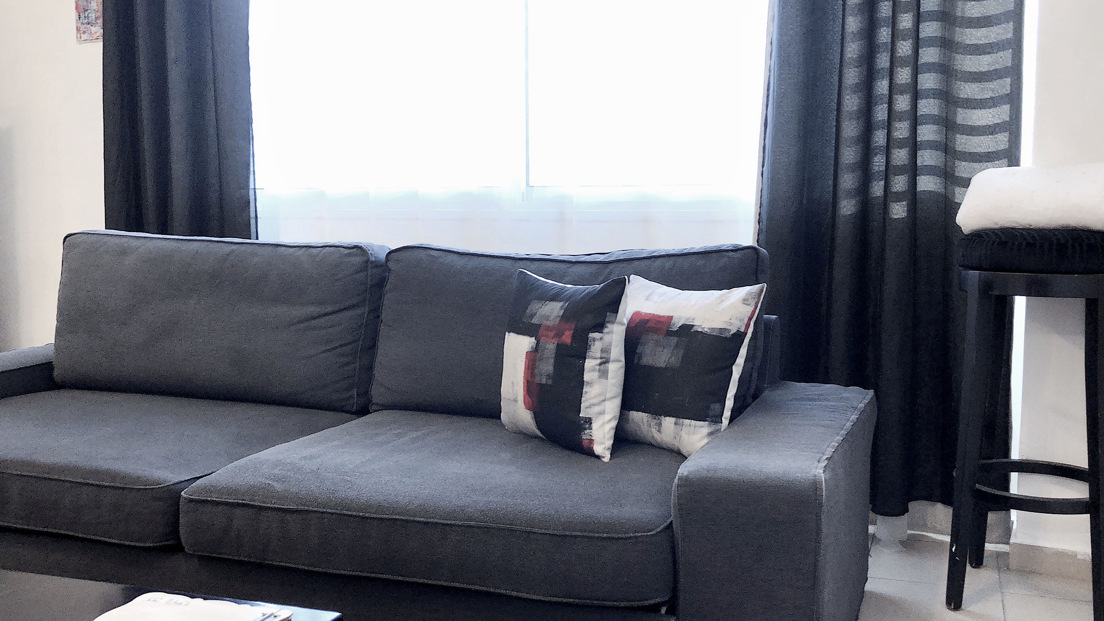 A grey sofa with a patchwork pillow, a black chair, and a window with curtains.