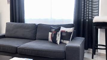 A grey sofa with a patchwork pillow, a black chair, and a window with curtains.