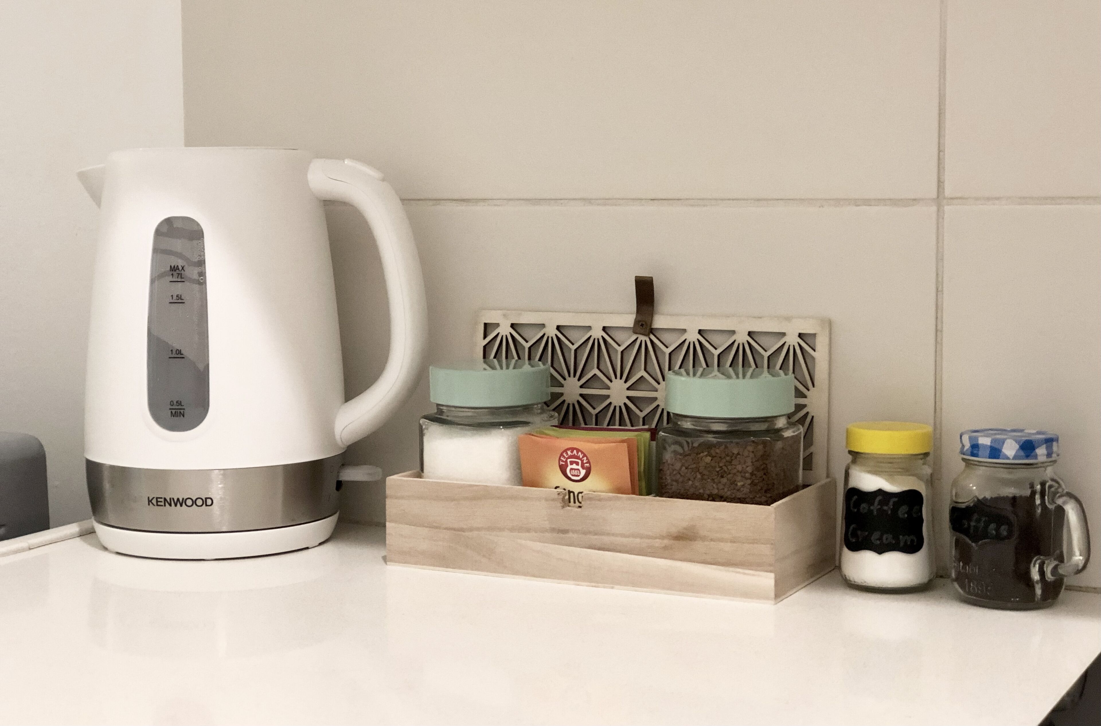 Una tetera blanca con marcas de medición, un organizador de especias de madera con frascos etiquetados como “Café Crema” y “Café”, y una pequeña caja con un logotipo.