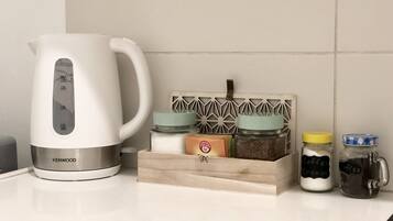 Una tetera blanca con marcas de medición, un organizador de especias de madera con frascos etiquetados como “Café Crema” y “Café”, y una pequeña caja con un logotipo.