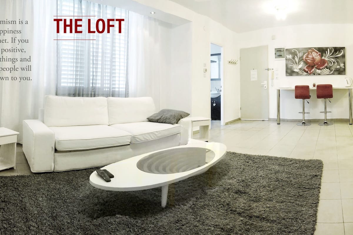 Um loft moderno com sofá branco, mesa lateral pequena, controle remoto sobre uma mesa de centro branca e tapete cinza.