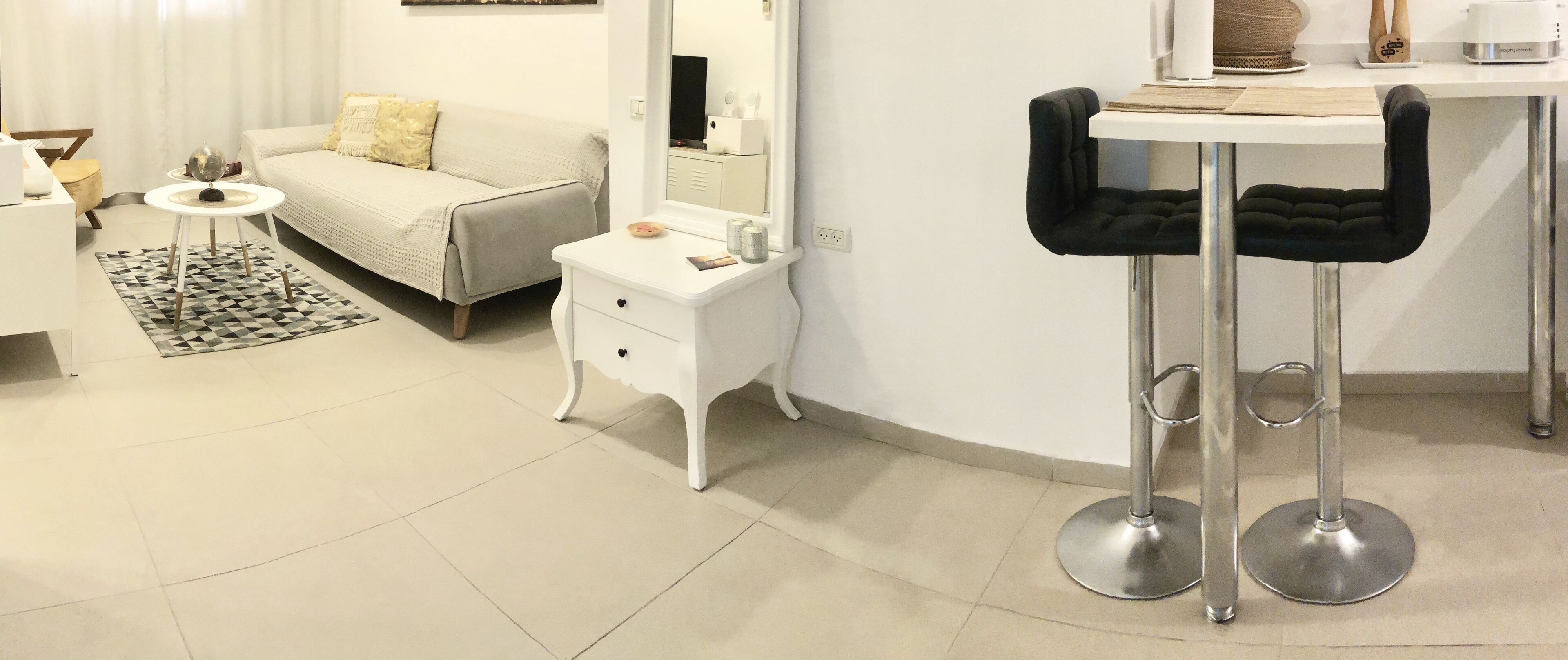 Une chambre d’hôtel moderne avec un canapé, une petite table ronde, une table de chevet blanche, un miroir et deux tabourets de bar noirs.