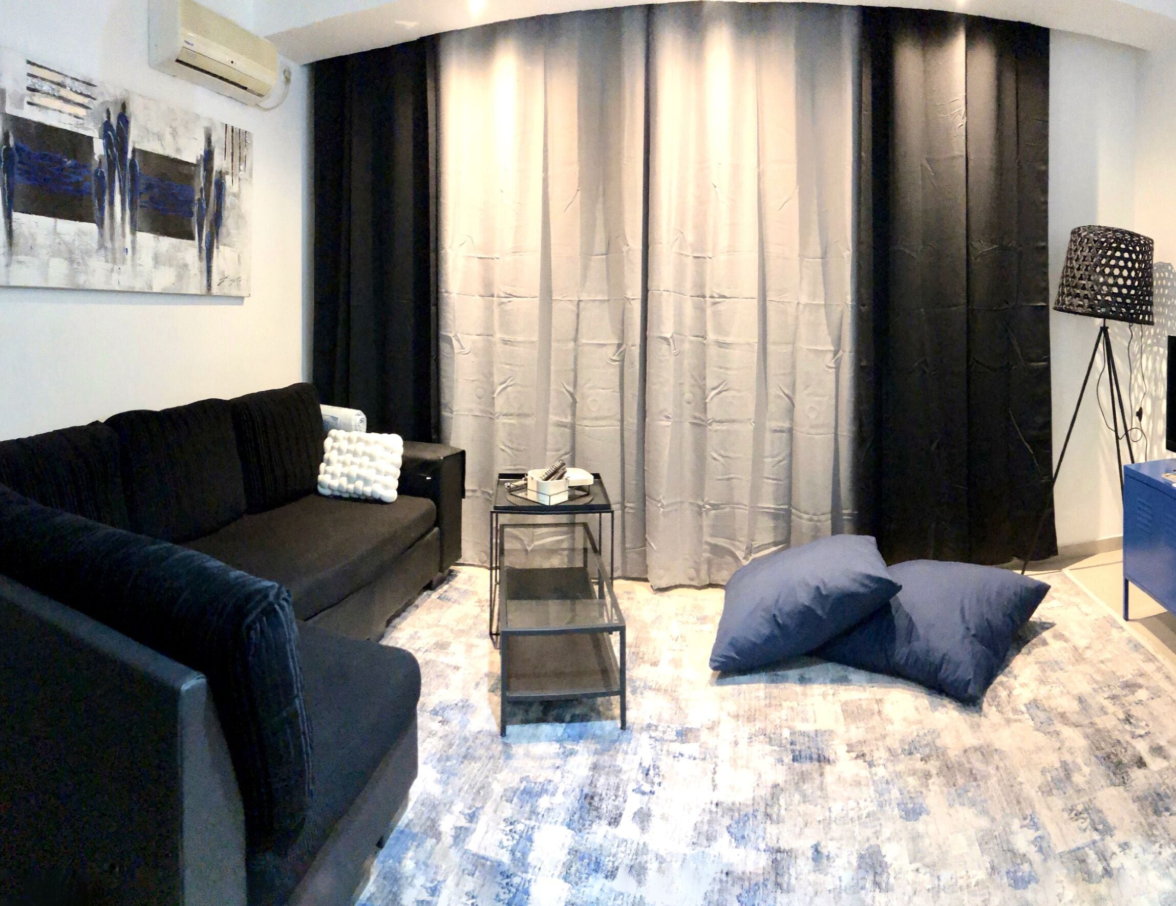A modern living room with a dark sofa, a glass coffee table, a blue pillow, and a floor lamp.