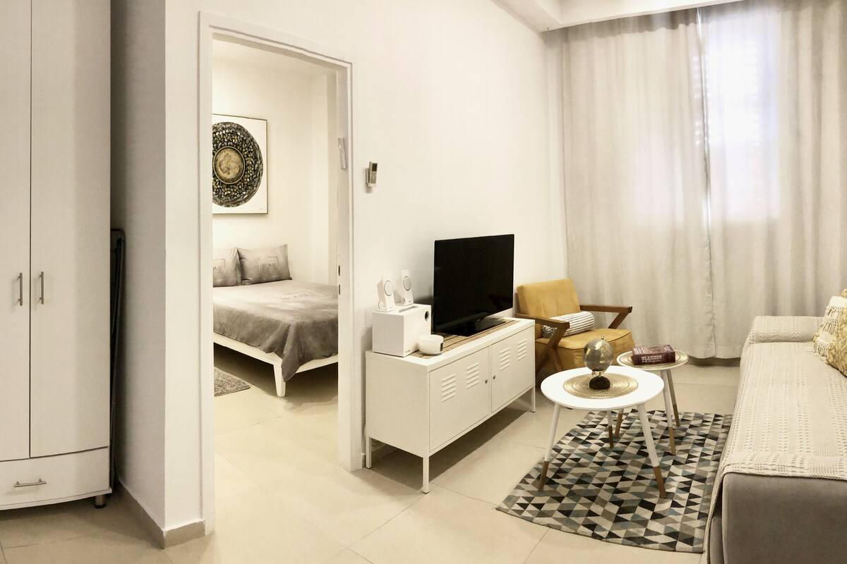 Quarto moderno e bem iluminado, com uma cama, TV, uma mesinha com alto-falantes e um sofá.