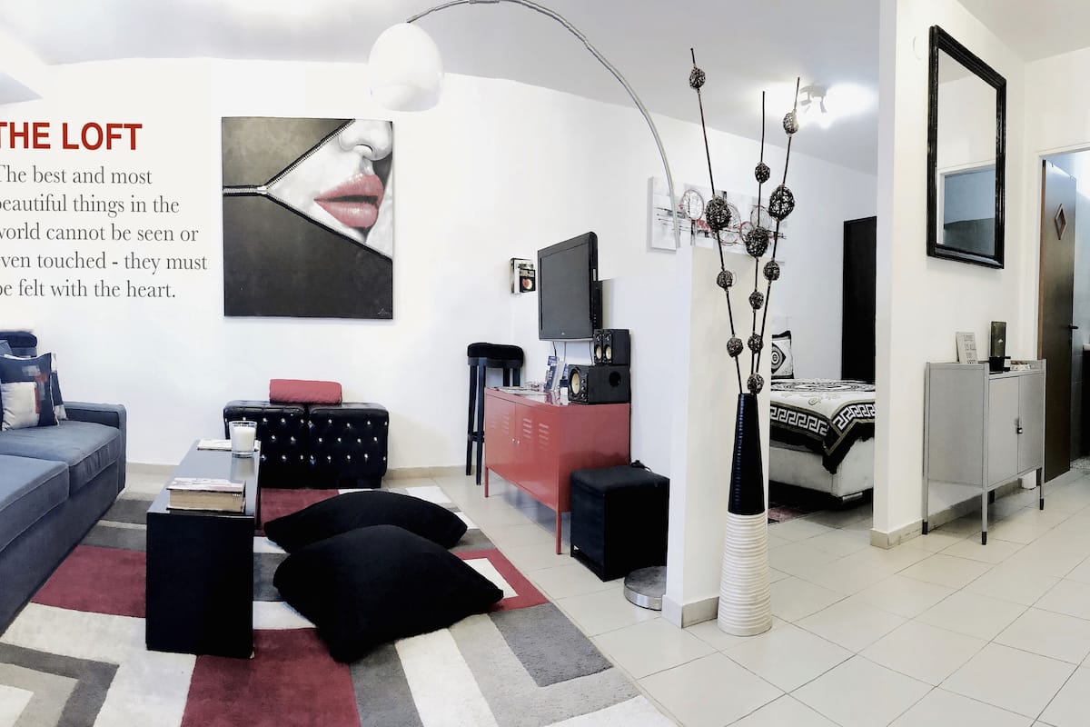 Uma sala de estar moderna com um sofá, mesa de centro e um armário vermelho. Há uma TV em um suporte e um vaso decorativo com ramos.