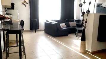 Un espacio moderno de sala con un sofá negro, una mesa de comedor con sillas y una cocina que incluye un refrigerador y armarios.