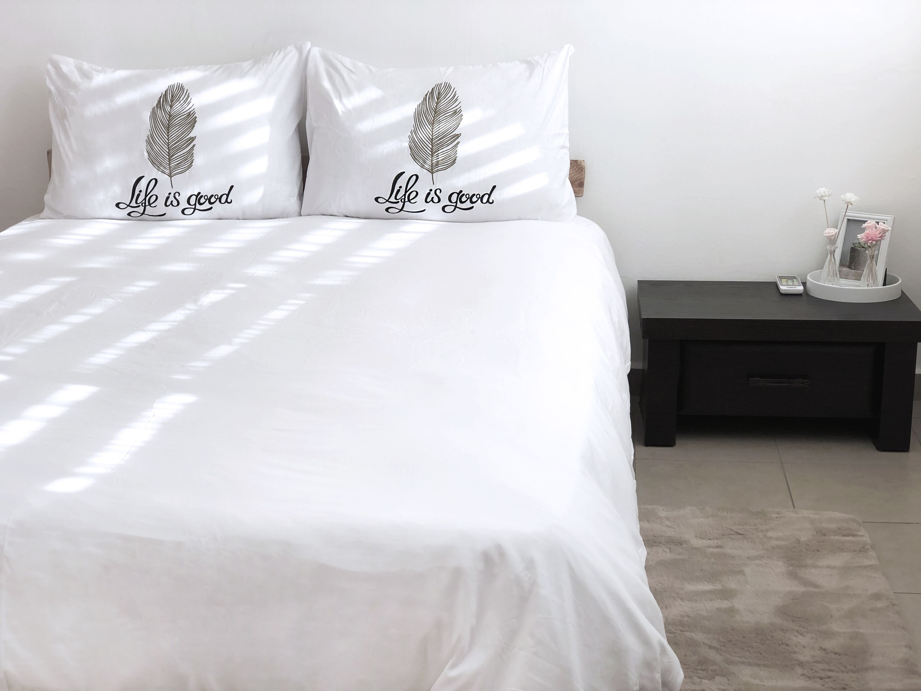 Una cama con ropa de cama blanca y dos almohadas con un diseño de plumas y el texto "Life is good".