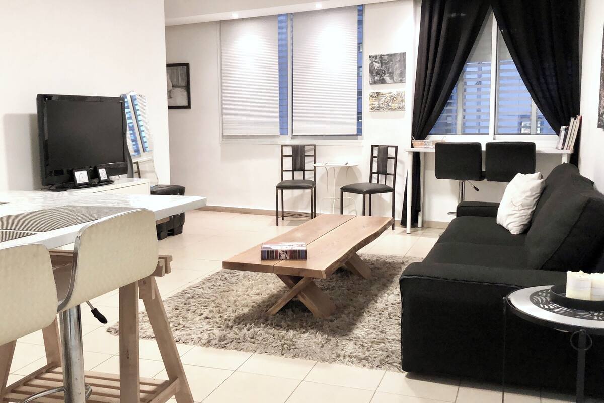 Uma sala de estar moderna com um sofá, uma mesa de centro e uma televisão. Há duas poltronas, uma mesinha e um tapete.