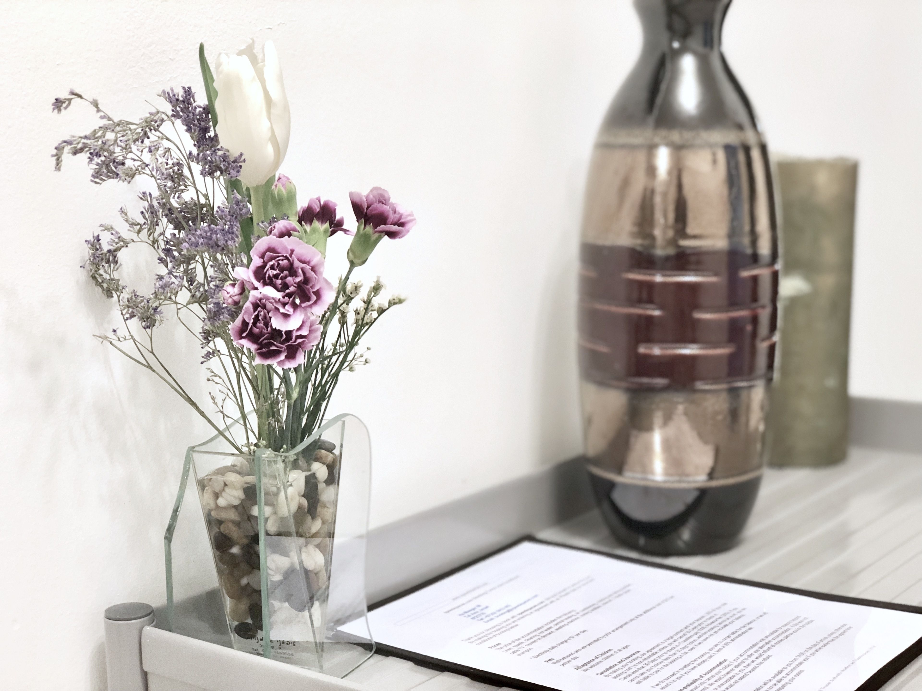 A clear glass vase with flowers and pebbles on a white surface next to a metal bottle and a document.