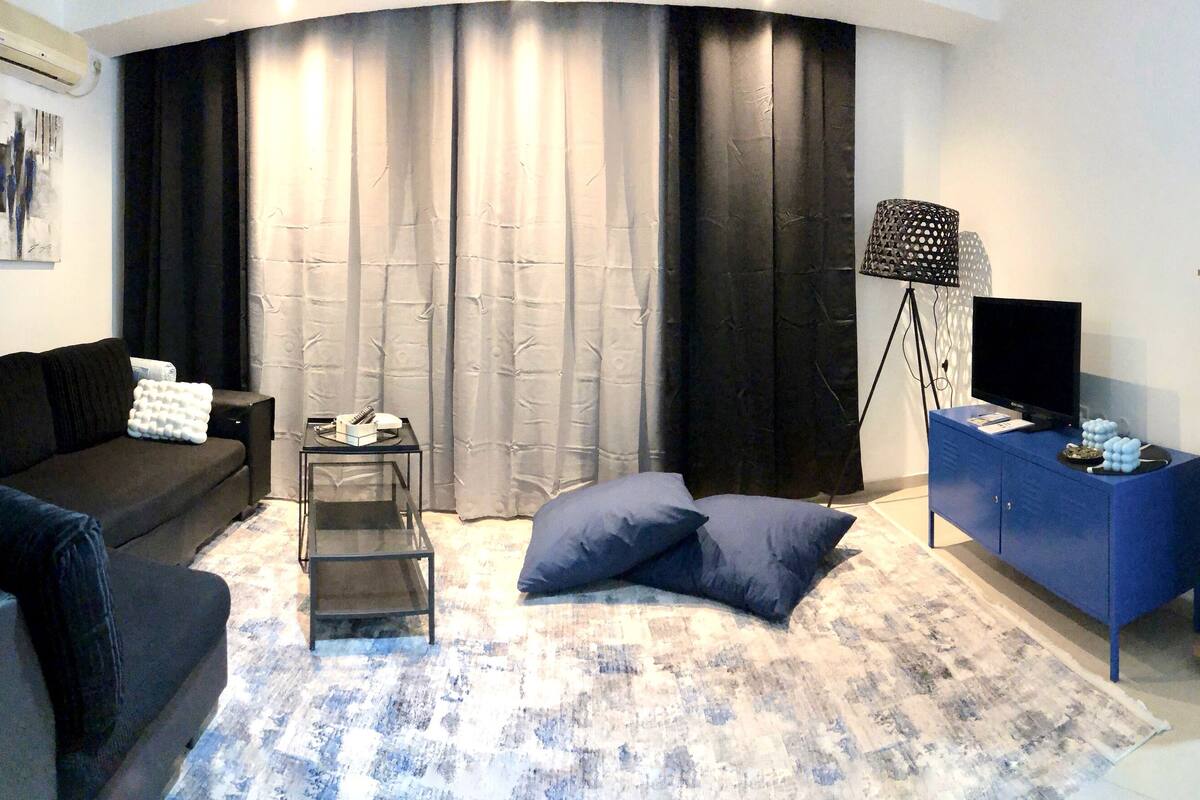 Uma sala de estar moderna com um sofá preto, uma pequena mesa lateral, um banquinho azul, um abajur e uma estante de livros.